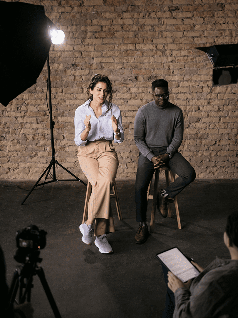 Two individuals seated on stools in a studio setting with photography equipment around.
